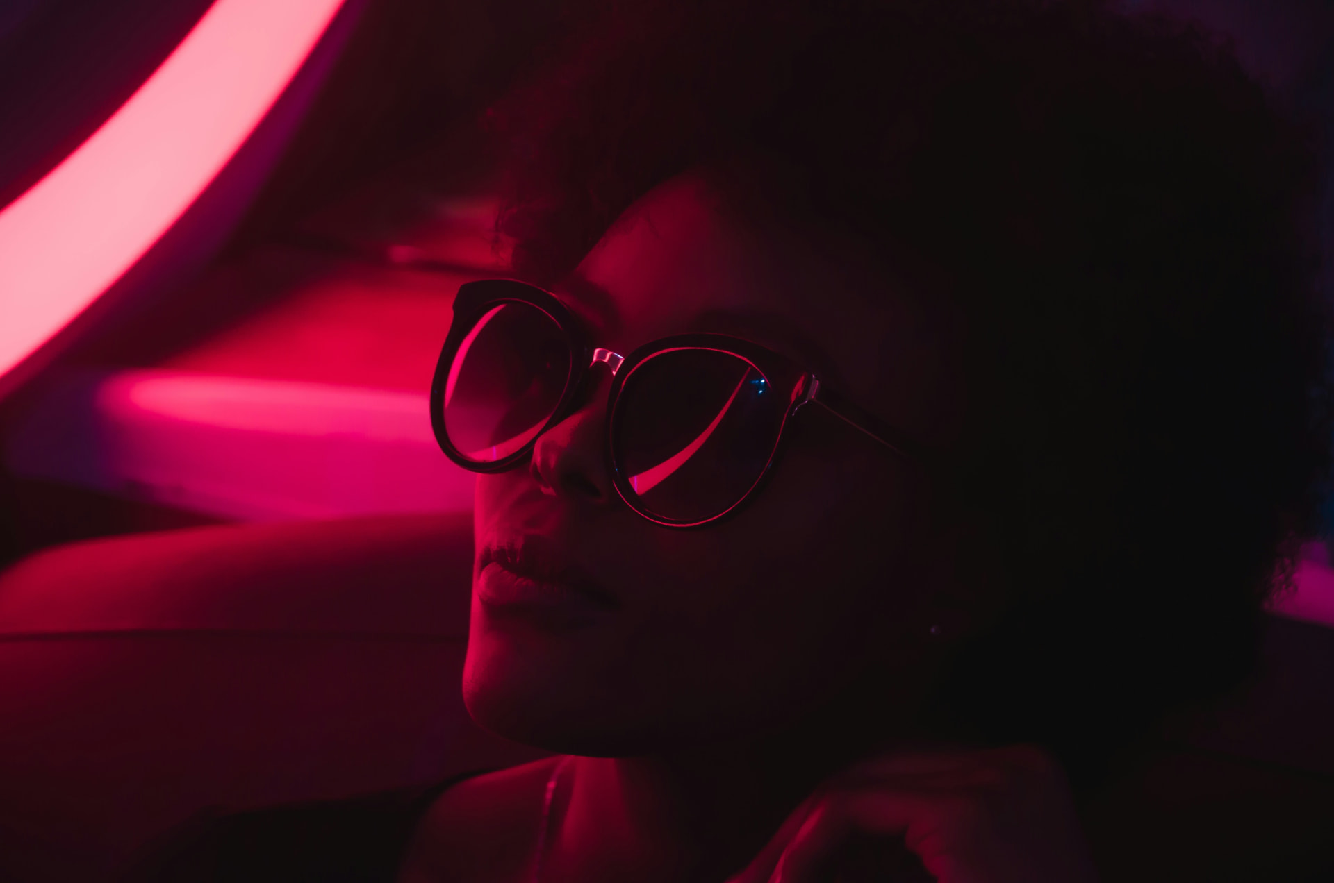 A woman in dark sunglasses lit by neon red and magenta light against heavy shadows
